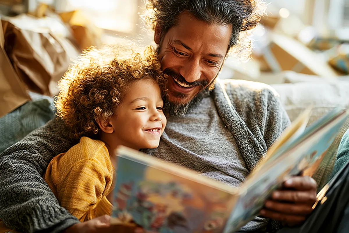A father and young child (around 3-5 years old) sitting together on a couch on what is clearly a celebration morning — wrapped gifts nearby, the child leaning against the father with clear affection. The father is holding a picture book open, looking at it with an expression of genuine surprise and delight. The child is watching his reaction. The atmosphere is warm, weekend-morning cozy. The gift clearly matters.