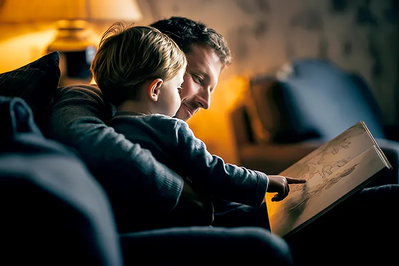 A father sitting in a large armchair in the early evening, a young child tucked against his side, both looking at an open picture book. The child's finger points at an illustrated character on the page. Warm lamp light. A relaxed, unhurried feeling — neither looking at anything except the book. Muted navy and amber tones. No faces fully visible. The focus is on their shared attention, the small hand reaching up to point, the father's arm around the child's shoulder.