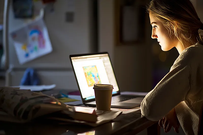 A parent at a kitchen table with a laptop open, looking focused but not panicked — a birthday invitation and a wrapped box visible nearby, a child's drawing on the fridge in the background. The laptop screen shows a design interface for a personalized book. A cup of coffee, a slightly scattered but functional workspace. The atmosphere is: someone who left it a bit late but has a plan. Late afternoon light. Competent calm.
