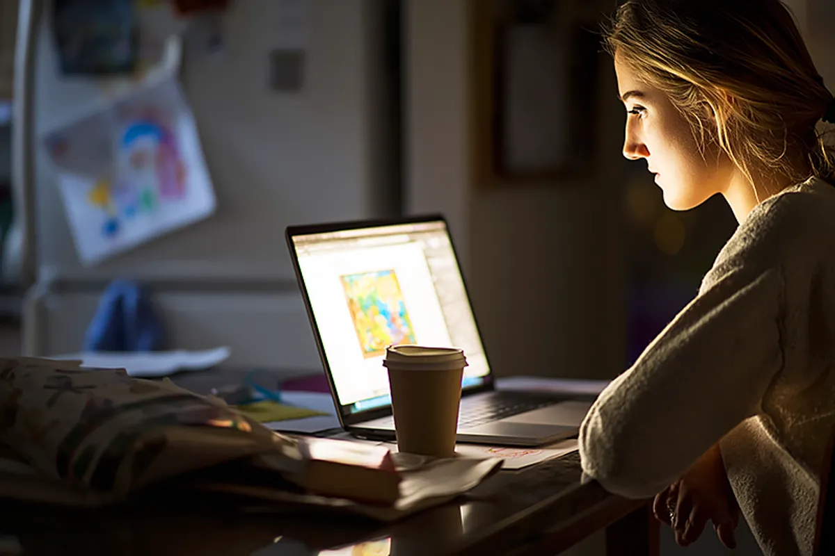 A parent at a kitchen table with a laptop open, looking focused but not panicked — a birthday invitation and a wrapped box visible nearby, a child's drawing on the fridge in the background. The laptop screen shows a design interface for a personalized book. A cup of coffee, a slightly scattered but functional workspace. The atmosphere is: someone who left it a bit late but has a plan. Late afternoon light. Competent calm.