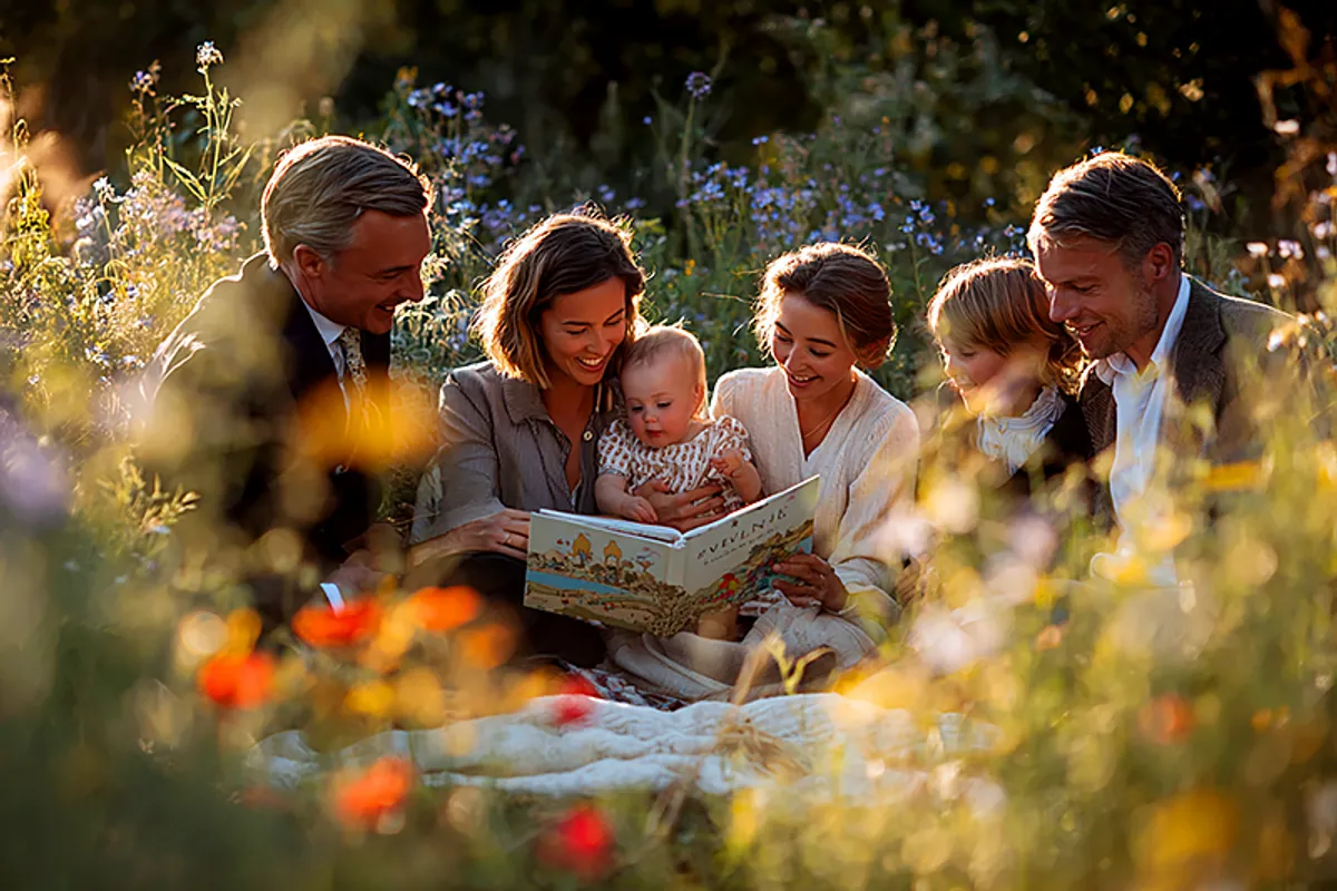 A garden naming ceremony scene: a small group of adults gathered in warm afternoon sunlight around a baby held by a parent. A beautifully illustrated personalized children's book sits open on a blanket in the foreground. No religious symbols. Wildflowers, natural linen textures, warm golden light. Painterly style, intimate and joyful.