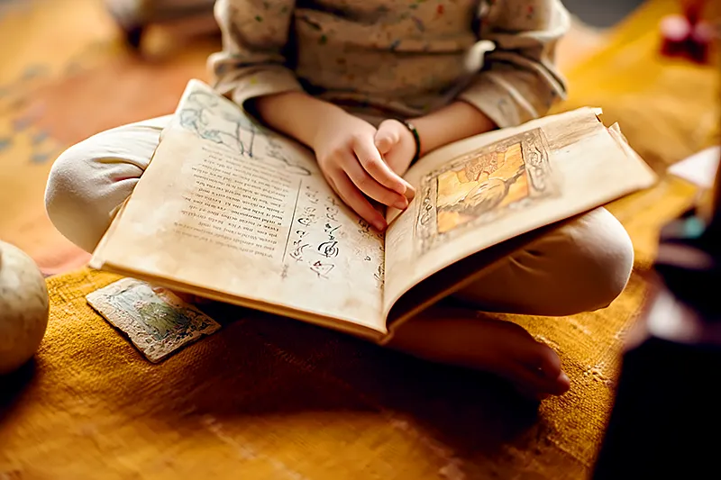 A child sitting cross-legged on a warm-toned rug, holding an open storybook. The visible page spread shows text in two languages side by side — one in English, one in elegant Mandarin characters — framing a glowing watercolor illustration of a child character who looks just like the reader. The scene feels cozy and culturally rich: a few objects nearby suggesting heritage (a small ceramic vase, a painted tile). Soft afternoon light, intimate and warm.