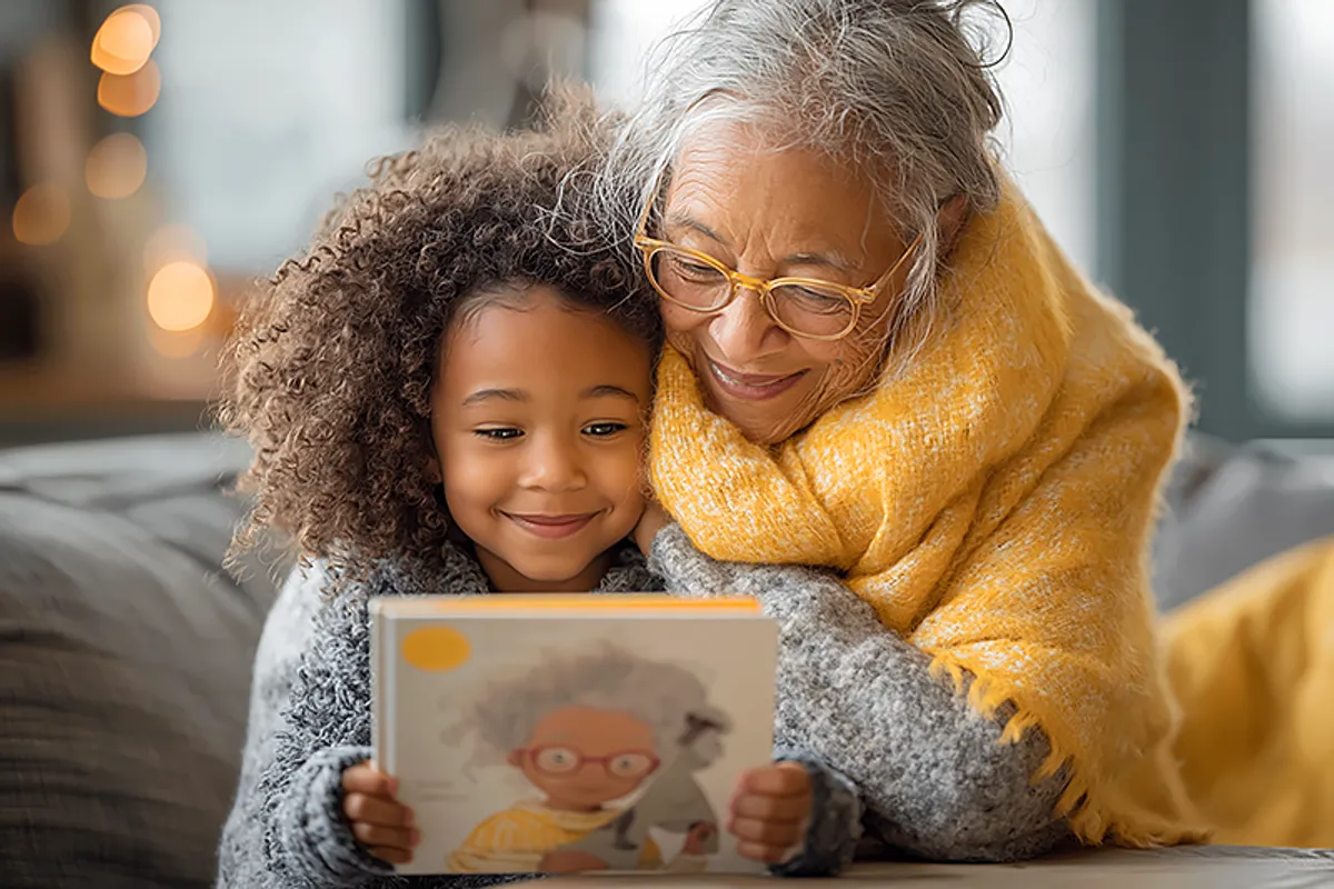 A grandmother — warm, clearly someone who arrived with intention and love — sitting with a young grandchild of around 3-5 on a sofa or at a kitchen table. The child has just opened a gift and is holding a picture book, staring at it with wide eyes and an expression of complete recognition. The grandmother has one arm around the child, watching their reaction with obvious, quiet delight. The book's cover shows an illustrated character who looks just like the child. Soft natural light through windows, the comfortable warmth of a well-loved home. A moment of being completely seen.