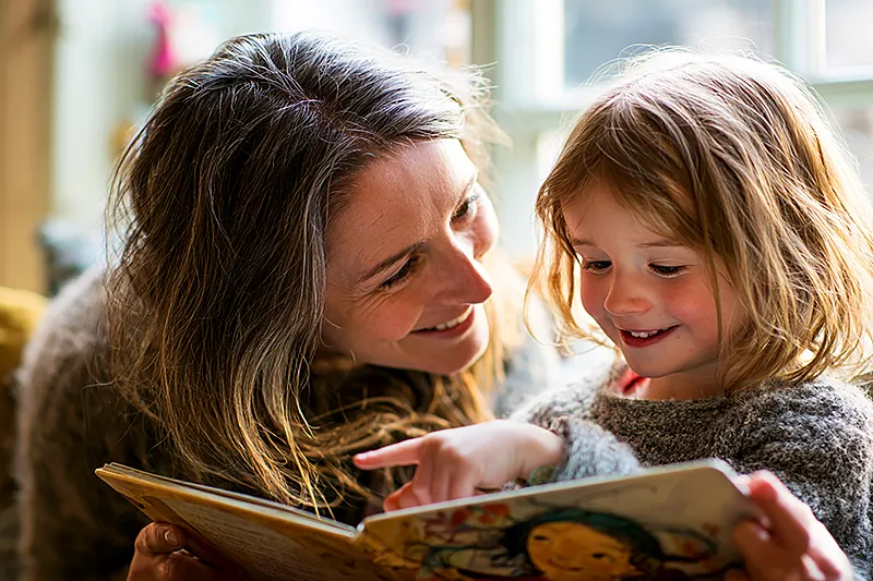 An Irish family scene — a parent and young child of around four or five sitting together in a warm, characterful home interior: a bright kitchen or cozy sitting room with the feel of a well-loved Irish house. The child holds a picture book open with both hands, pointing excitedly at an illustrated character who looks just like them. The parent leans in, face lit with delight. Natural light from a window, the comfortable warmth of family life. A sense of home.