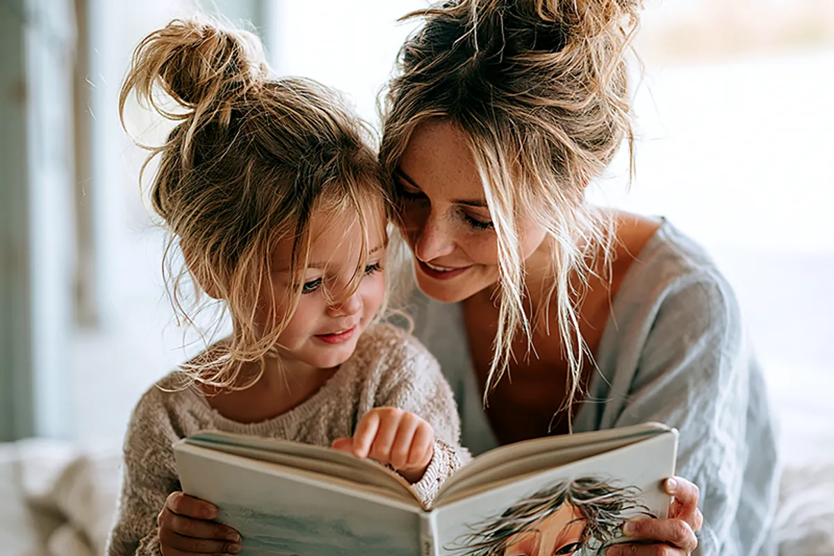 A New Zealand family scene — a parent and child of around four or five, sitting together outside or in a bright, light-filled room with the quality of a New Zealand summer afternoon. The child is holding a picture book open with both hands, pointing excitedly at an illustrated character who looks just like them. The parent is leaning in with delight. The atmosphere is warm, relaxed, and distinctly Southern Hemisphere — bright light, open space, the feel of a long summer day. A loved and lived-in family home.