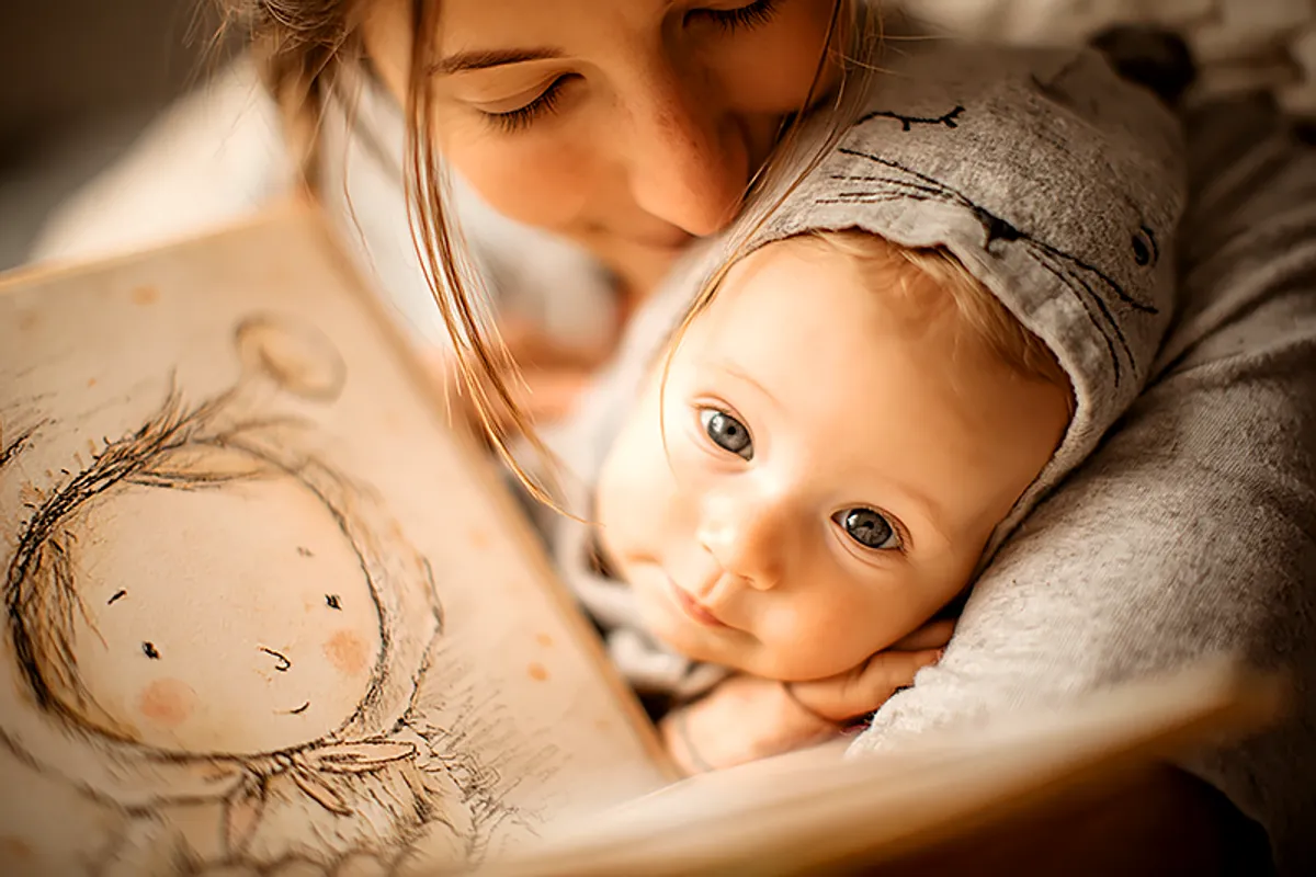 A parent holding an infant — a rainbow baby — with a picture book open nearby. The book shows a beautifully illustrated character that resembles the baby: soft features, the same expression. The parent is looking at the baby, not the book, with an expression of profound tenderness and something that is almost disbelief — in the good sense. The atmosphere is quiet, intimate, and full of warm light. A sense of arrival, of something waited for and now real.