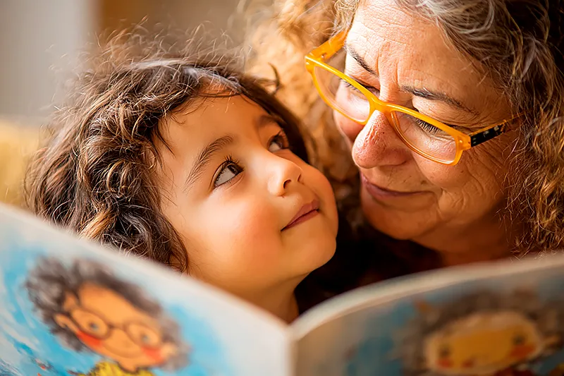 A parent and a young child of about four or five, close together in a warm and comfortable reading space. The child is listening intently, face tilted upward toward the parent's voice, while the parent reads from a picture book whose illustrated pages are visible to us — showing a character who looks exactly like the child. The parent's expression is one of total engagement, reading with care. Natural warm light. The image conveys the intimacy of being read to, the child receiving the story through the parent's voice rather than their own eyes.