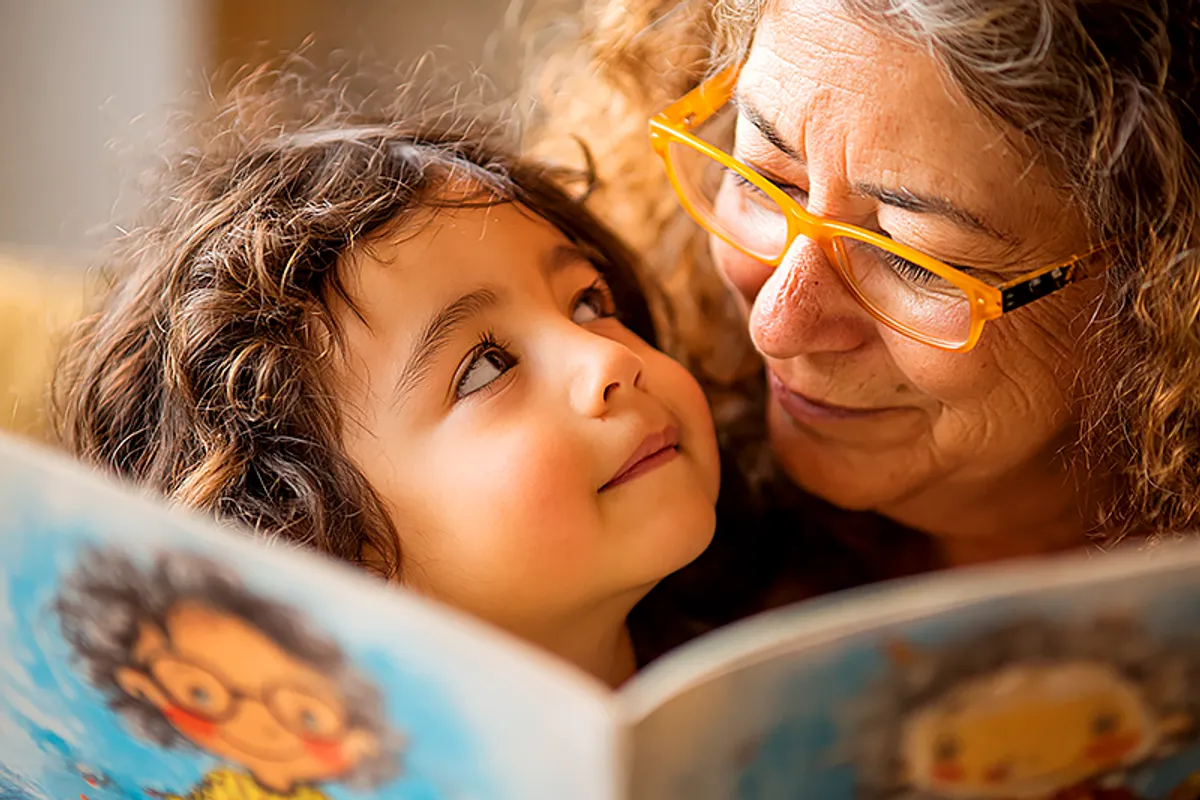 A parent and a young child of about four or five, close together in a warm and comfortable reading space. The child is listening intently, face tilted upward toward the parent's voice, while the parent reads from a picture book whose illustrated pages are visible to us — showing a character who looks exactly like the child. The parent's expression is one of total engagement, reading with care. Natural warm light. The image conveys the intimacy of being read to, the child receiving the story through the parent's voice rather than their own eyes.
