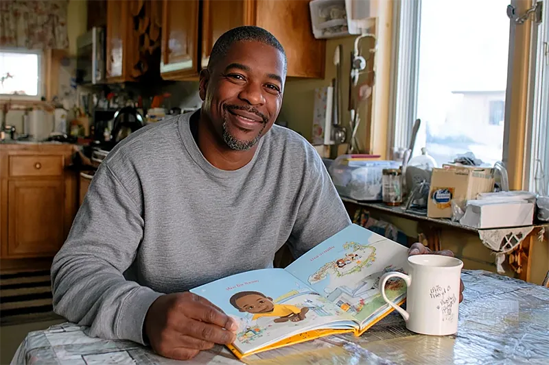 A father sitting at a kitchen table on a quiet Sunday morning, holding a small hardcover children's book. The book is open; the illustrated cover is visible — clearly a personalized book, with a child's likeness on the cover. On the table: a coffee mug, a Sunday paper folded back, the ordinary texture of a family morning. His expression is private and genuine — not performed delight, but the look of someone holding something that actually matters. Warm morning window light. No party, no ribbons.