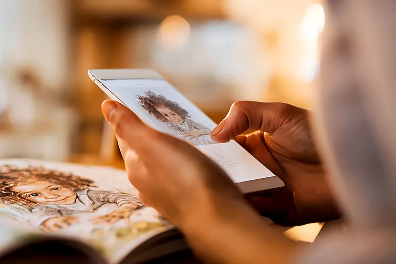 Close-up of a parent's hands holding a smartphone showing a child's photo, with a personalized children's book open on the table beside it showing the same child illustrated in a watercolor storybook style. Warm kitchen light, shallow depth of field. The phone screen shows an upload interface. Cozy, safe, domestic atmosphere. Soft focus background.