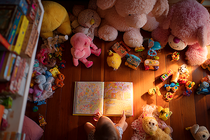 A child's playroom seen from above, overflowing with colorful toys and stuffed animals, but in the center of the floor sits a single open personalized storybook glowing with warm light. The child's hand reaches toward it, ignoring everything else. Painterly bird's eye perspective, warm palette, contrast between abundance and meaning.