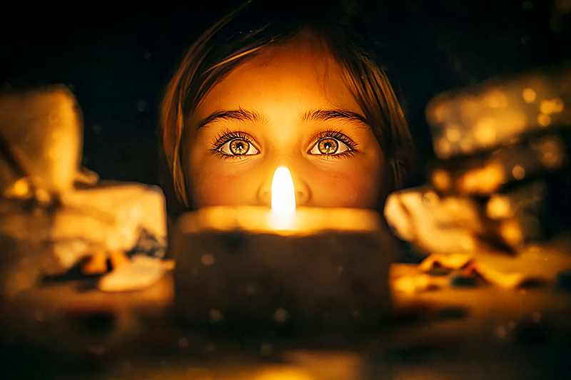 A single birthday candle glowing on a small cake, reflected in the wide eyes of a child looking at it in wonder. Around the cake, blurred in bokeh, are wrapped presents and scattered confetti. The focus is entirely on the child's face and the candle flame. Painterly style, warm golden light, intimate and reverent.